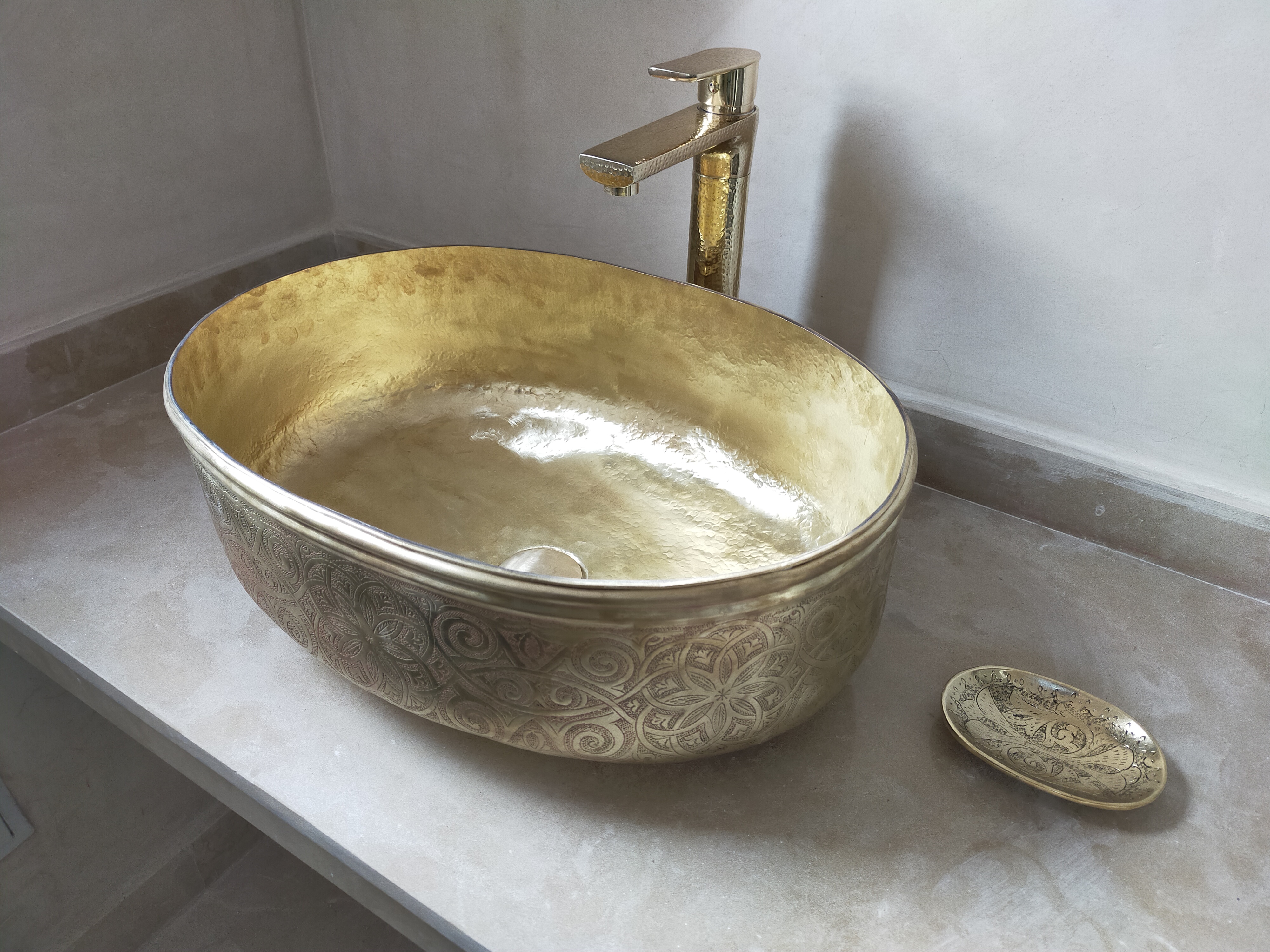 Lavabo en cuivre travaillé artisanalement, orné de motifs détaillés, accompagné d’une robinetterie dorée et d’un plateau décoratif sur un comptoir en marbre clair.