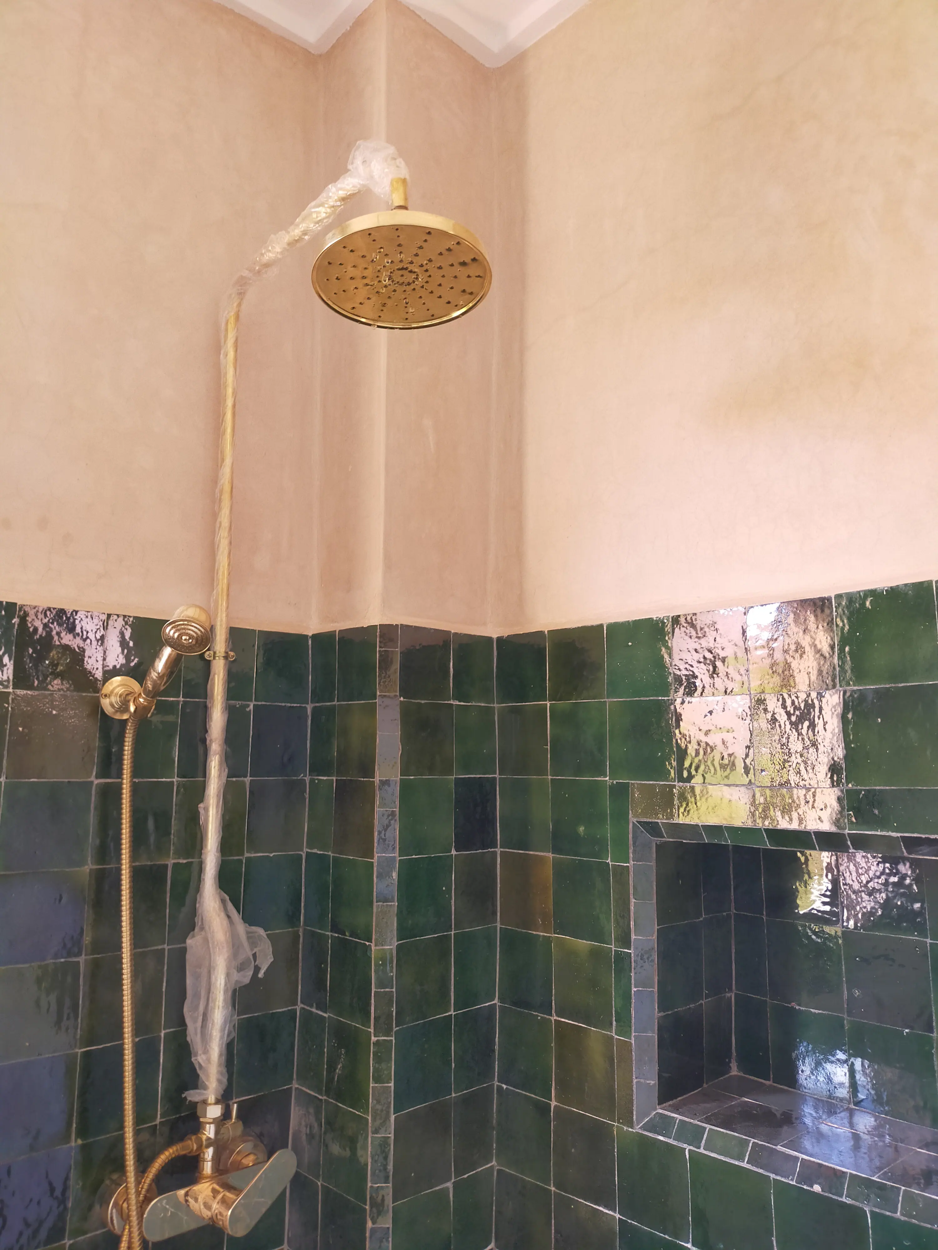 Un hammam marocain moderne aux murs en carreaux de faïence verts, doté d’une douche murale avec robinetterie dorée et un bassin intégré pour une expérience de détente authentique.