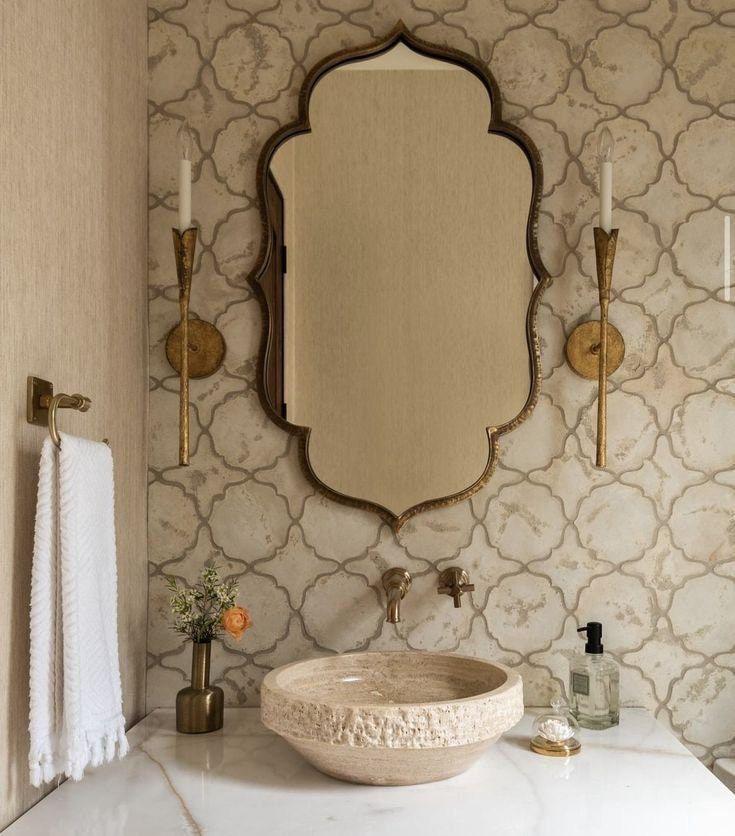 Salle de bain raffinée aux murs en carreaux de faïence beige ornés, dotée d’un miroir à cadre doré, d’un lavabo en pierre naturelle et de lampes murales décoratives.