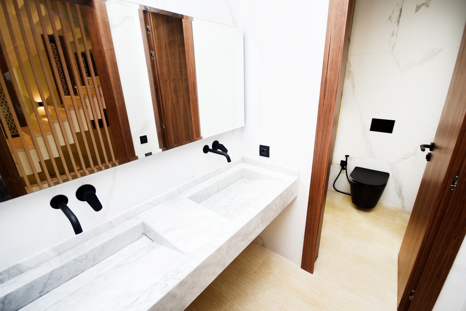 Salle de bain contemporaine aux murs blancs, dotée d’un double lavabo en marbre intégré dans un comptoir long, de robinetterie noire et d’un WC suspendu noir
