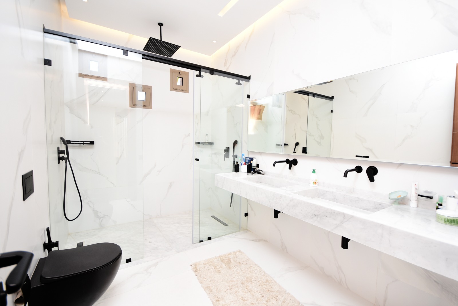 Salle de bain contemporaine aux murs en marbre blanc veiné, dotée d’une douche à porte coulissante en verre transparent, d’un meuble suspendu avec double vasque et robinets noirs, ainsi que d’un WC design noir. L’éclairage intégré au plafond apporte une touche de modernité.