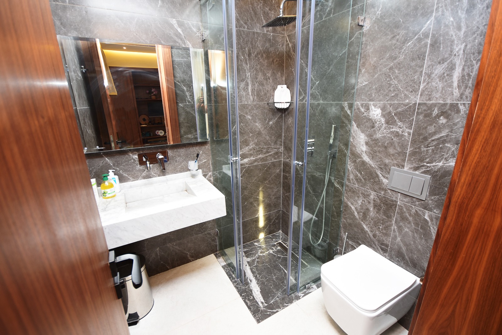Salle de bain moderne aux murs en marbre noir veiné, dotée d’une douche à porte coulissante en verre transparent, d’un meuble sous vasque blanc, d’un WC suspendu et d’un éclairage ambiant doré au plafond.