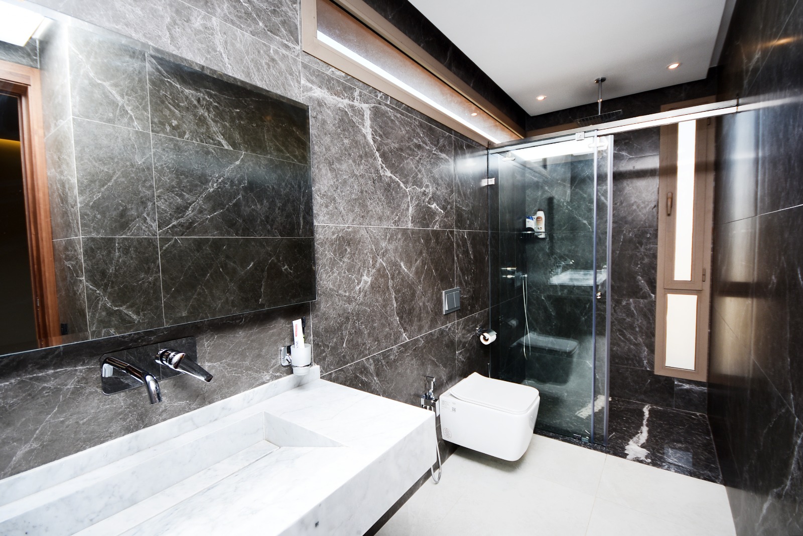 Salle de bain moderne aux murs en marbre noir veiné, dotée d’une douche à porte coulissante en verre transparent, d’un meuble sous vasque en marbre blanc et d’un WC suspendu. L’éclairage LED au plafond apporte une touche de sophistication