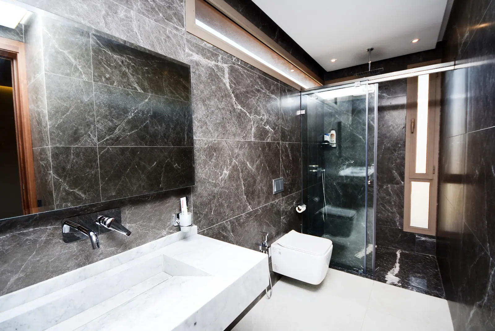 Salle de bain moderne aux murs en marbre noir veiné, dotée d’une douche à porte coulissante en verre transparent, d’un meuble sous vasque en marbre blanc et d’un WC suspendu. L’éclairage LED au plafond apporte une touche de sophistication