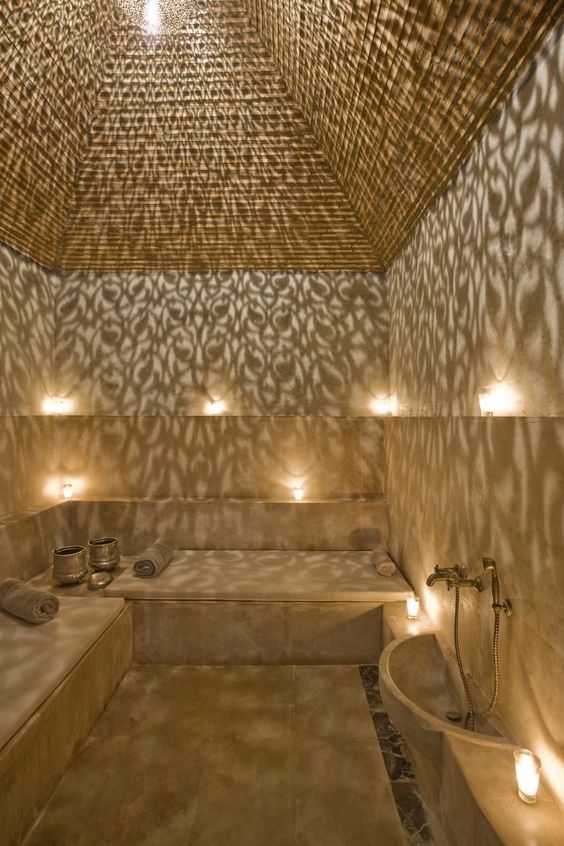Un hammam marocain luxueux aux murs texturés, doté de bancs en marbre beige, d’une lampe suspendue avec motifs qui projette des ombres décoratives, et d’un éclairage doux pour une ambiance relaxante