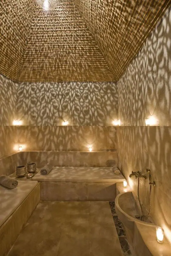 Un hammam marocain luxueux aux murs texturés, doté de bancs en marbre beige, d’une lampe suspendue avec motifs qui projette des ombres décoratives, et d’un éclairage doux pour une ambiance relaxante